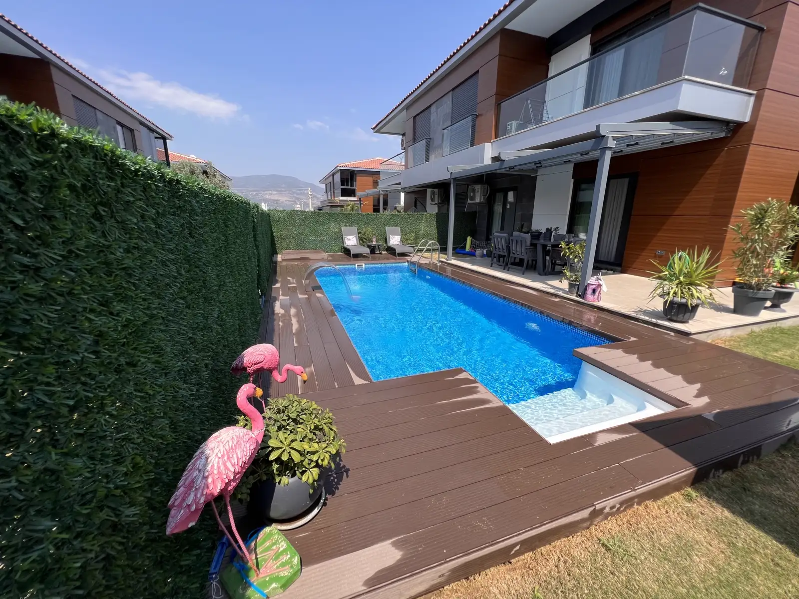 Dar villa bahçesinde ahşap deck üzerinde uzun dikdörtgen havuz, bir ucunda geniş basamak/oturma nişi ve paslanmaz merdiven